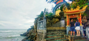 Daytime view of the Dinh Cau temple gate and shrine on a rocky outcrop above the sea, with two visitors at the altar