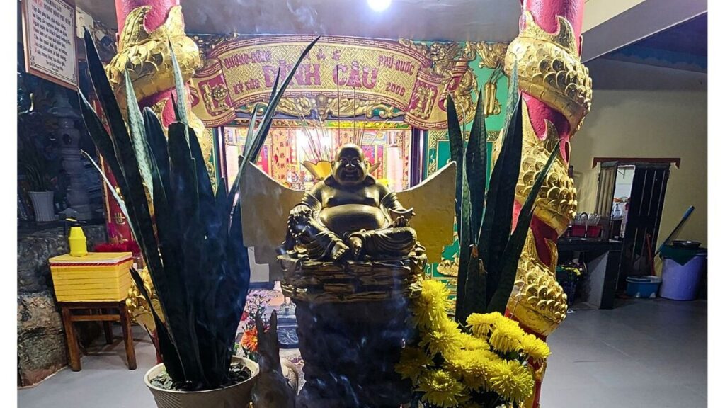 Golden laughing Buddha statue inside the Dinh Cau shrine, framed by red and gold dragon pillars and yellow chrysanthemum offerings