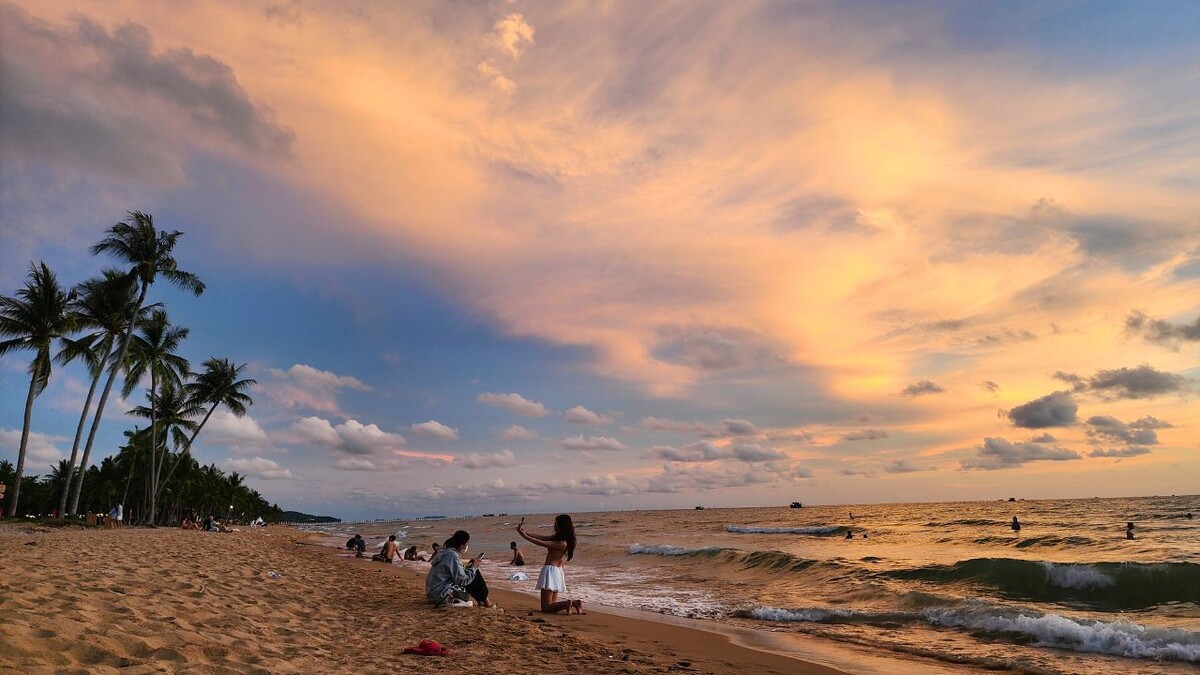 People on the beach taking photos under a dramatic orange sunset sky with palm trees at Phu Quoc Marina